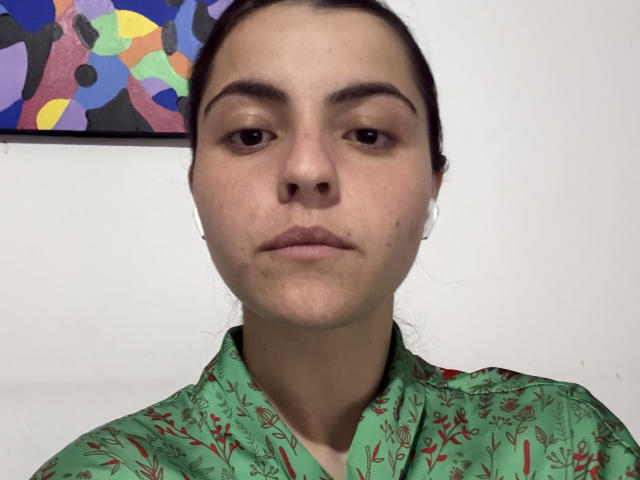 MarchelleFaubert tem uma expressão neutra, pele clara, olhos castanho-escuros e está vestindo uma camisa verde com estampas florais vermelhas. Seu cabelo castanho está preso para trás..