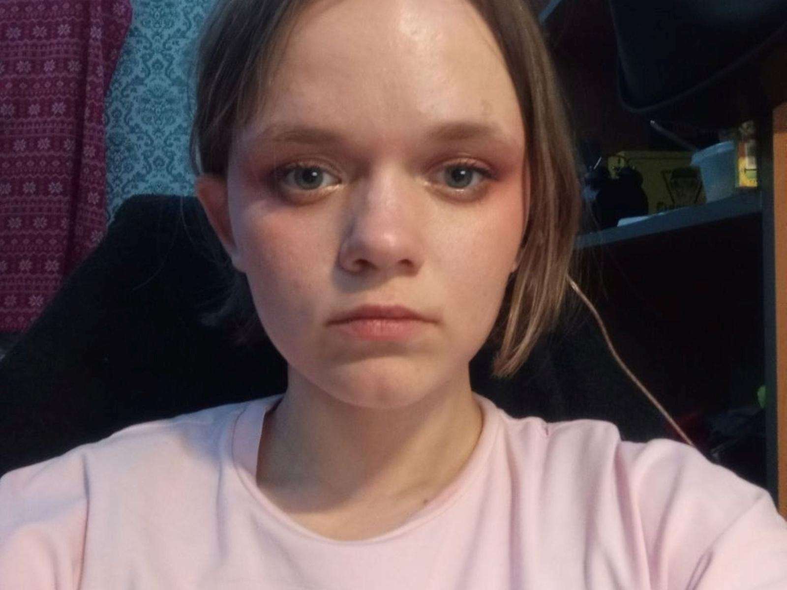 AngeliaAmstutz has a tired, puffy face with blue eyes, wearing a pink shirt, looking slightly to the side with a serious expression, and brown hair.
