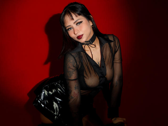 SophiaBlakes with black mesh top, showing her tits through the fabric, wearing a shiny black skirt, red background, dark red lipstick, and black hair with bangs.
