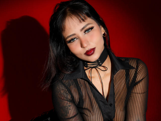 SophiaBlakes met een zwarte doorschijnende top, rode lipstick en een zwarte choker, verleidelijk kijkend naar de camera, waarbij haar borsten zichtbaar zijn door de stof, op een rode achtergrond..
