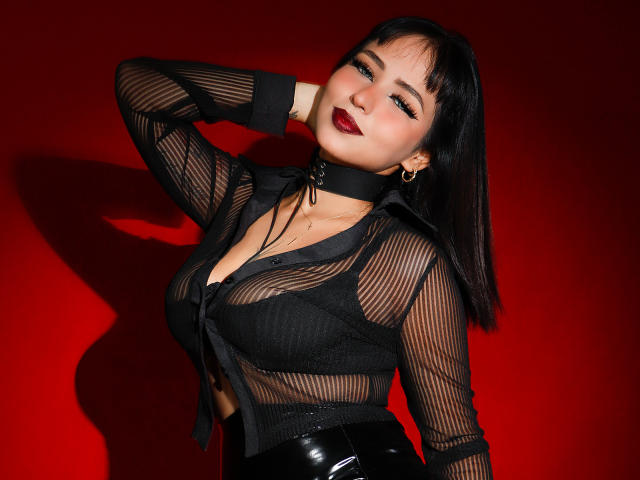 Sophia, vêtue d'un haut noir transparent qui révèle ses seins, portant un collier noir et un pantalon noir brillant, sur un fond rouge, avec du rouge à lèvres foncé et des cheveux noirs..