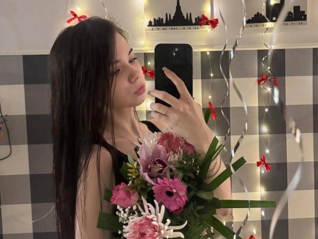 AngelicMcentire, com longos cabelos pretos, segurando um buquê de flores rosa e brancas, tirando uma selfie no espelho com um celular preto, luzes de fada e fitas vermelhas ao fundo..