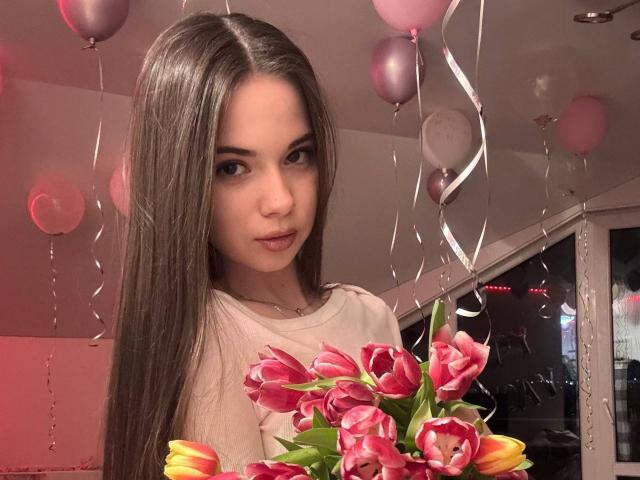 AngelicMcentire con lunghi capelli castani, occhi di colore rosa e rossetto rosa, che tiene un bouquet di tulipani rosa e gialli, circondata da palloncini rosa e nastri argentati..