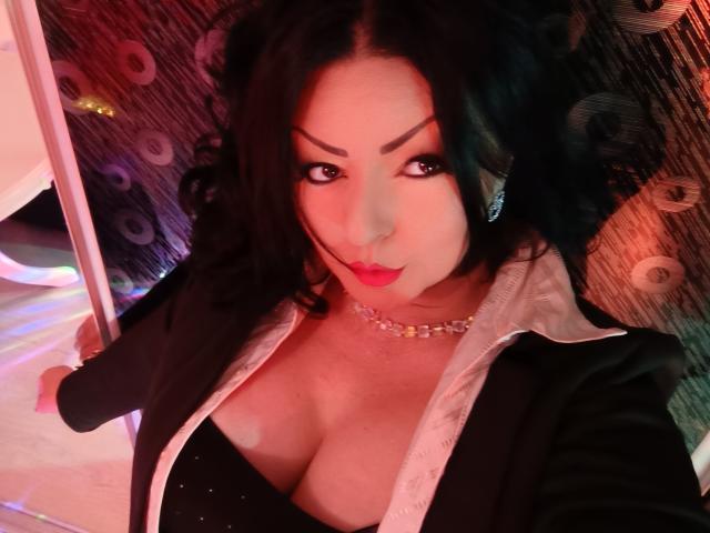 AmberSunshine has black curly hair, bold eyebrows, red lips, and is wearing a black jacket with a white lining, showing her large tits in a low-cut top.
