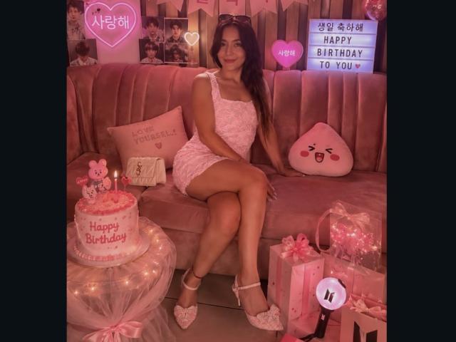 MindyLove met een gemiddelde borstmaat, in een roze kanten jurk, zittend op een roze bank, benen gekruist, witte pumps in de vorm van een hart, een verjaardagstaart met de tekst "Fijne Verjaardag" erop, cadeaus eromheen..