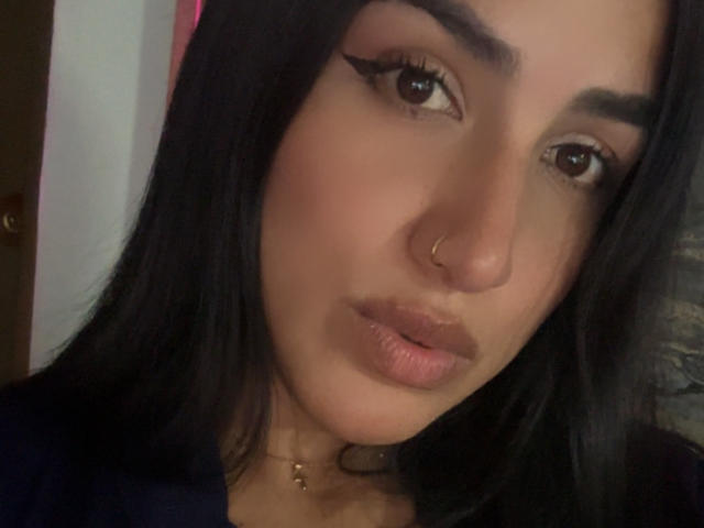 Alana Harris tem olhos castanho-escuros, cabelo preto, um piercing no nariz e um colar de ouro, com lábios rosados e maquiagem discreta, olhando diretamente para a câmera..