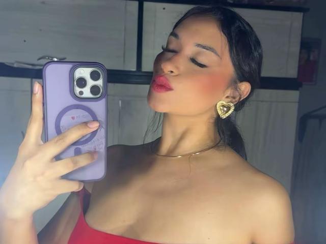 Adalina Vera mit sinnlich geschwungenen, roten Lippen, trägt ein rotes Top ohne Träger, goldene herzförmige Ohrringe und eine violette Handyhülle und macht ein Selfie mit geschlossenen Augen. Ihre Brüste sind leicht sichtbar..