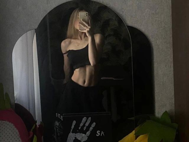 Selfie allo specchio di una ragazza attraente, con capelli biondi, top nero con una spalla scoperta e gonna nera, che mostra addominali scolpiti e tiene in mano il telefono, coprendo il viso. Il riflesso mostra uno specchio nero con impronte di mani..