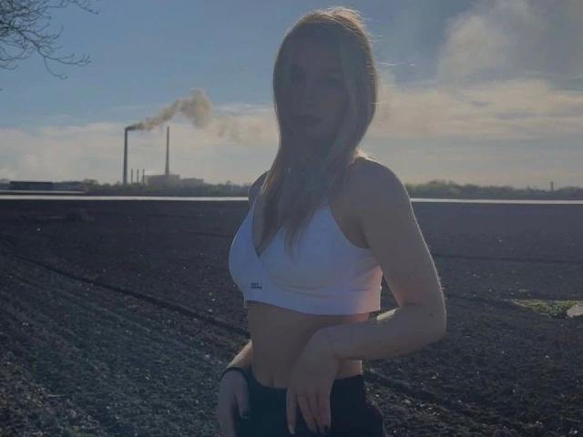 Une femme, probablement une "Stasi-girl", avec de longs cheveux blonds, un débardeur de sport blanc laissant entrevoir ses seins, et un pantalon noir, debout au soleil, les mains sur les hanches, avec une cheminée en arrière-plan et un ciel bleu clair..