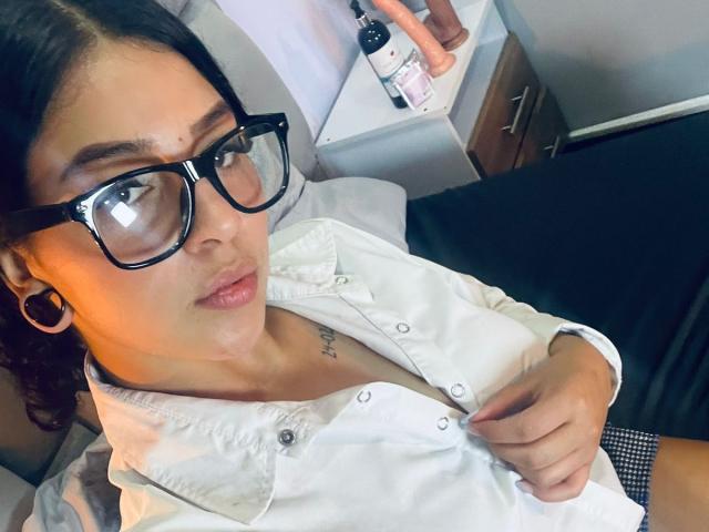 LonxEva with dark hair, glasses, white button-up shirt, and visible tattoo, looking at camera, hand on shirt, black ear gauge, dildo on table in background.