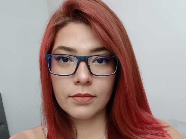LunaMedel hat rotes Haar, eine blaue Brille und ein ernstes Gesicht mit dezentem Make-up, darunter helfrosa Lippen und helle Haut. Ihr Hintern ist auf dem Bild nicht zu sehen..