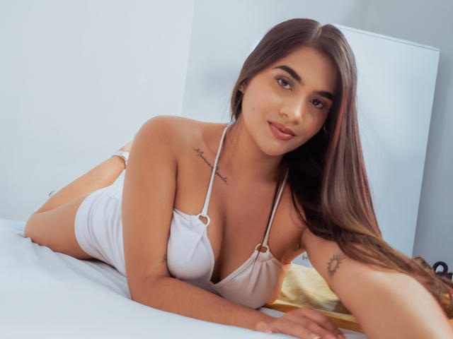 Isis Ferrer, met lang bruin haar, een gebruinde huid en kleine tatoeages, ligt op een bed in een witte haltertop, waarbij haar borsten zichtbaar zijn, en glimlacht naar de camera..