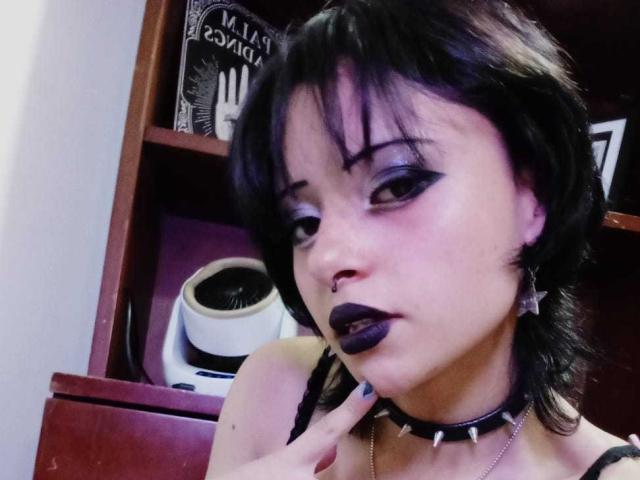 ViolettClay with black hair, purple eyeshadow, dark lipstick, nose piercing, spiked choker, and finger to mouth, showing off her tits and looking seductive.