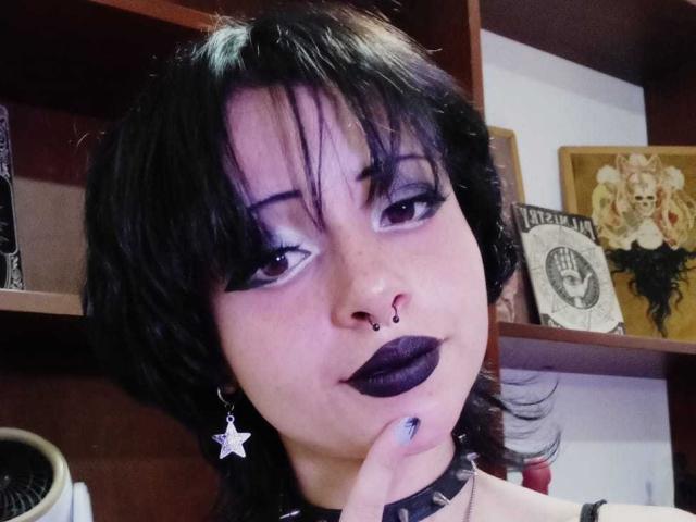 ViolettClay with black hair, purple eyeshadow, dark lipstick, nose ring, star earrings, choker, and finger touching her mouth, showing off her ass with her head tilted slightly.
