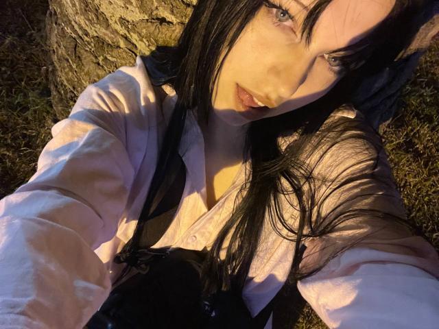 AmaiRin with long black hair, blue eyes, and pink shirt, lying outdoors, wearing black suspenders, showing cleavage, holding camera, sunlight on face, grass and tree in background.