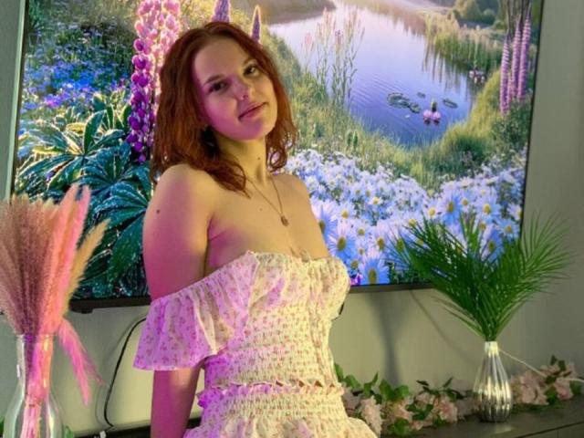 ChayaParrett with red hair, wearing a pink floral off-shoulder dress, standing in front of a colorful flower painting, holding a pink pampas grass vase on the right.