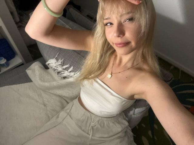 DonitaDunn with blonde hair, white strapless top, and beige pants, sitting on a gray couch, smiling, hand in hair, green bracelet, and a gold necklace.