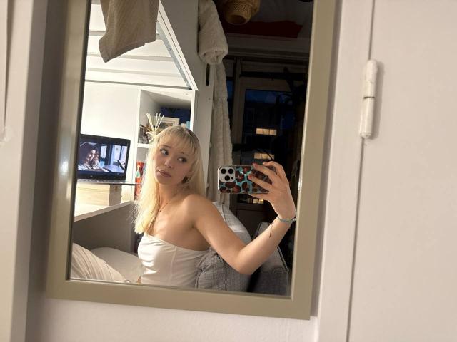 DonitaDunn with blonde hair, small tits, and a white strapless top, taking a mirror selfie with a phone in a dimly lit room. Reflection shows a TV in background.