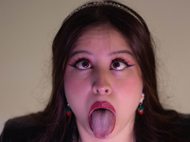 Het gezicht van Melani Becker, met grote ogen, donkere eyeliner en rode lippenstift, haar tong volledig uitgestoken, waardoor speeksel zichtbaar is. Ze heeft bruin haar, een hoofdband en rode oorbellen..