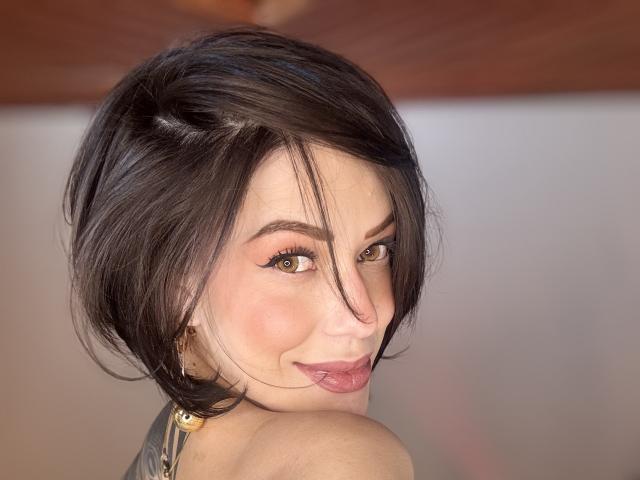 HannaBorbon, con cabello negro corto, ojos marrones y lápiz labial rosa, sacando la lengua, mostrando su hombro tatuado, y usando un collar negro..