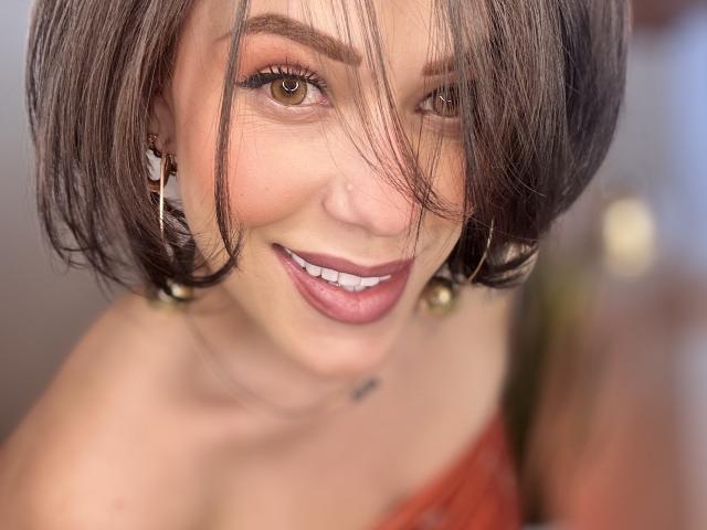 HannaBorbon, com cabelo castanho curto, sorrindo, olhos castanhos, batom rosa, usando brincos de ouro, um vestido vermelho e um colar pequeno, olhando para a câmera. Seus seios estão parcialmente visíveis..