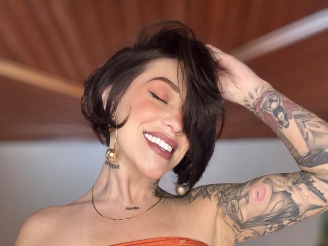 HannaBorbon con i capelli neri corti, che sorride, con gli occhi chiusi, una mano tra i capelli, che indossa rossetto rosso, orecchini di perle, un top senza spalline, tatuaggi sul braccio e sul collo, sfondo con soffitto in legno..