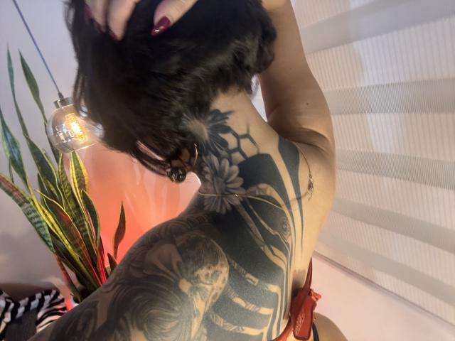 HannaBorbon, com cabelo preto, segurando a cabeça, exibindo uma grande tatuagem na lateral e nas costas, com flores e um rosto esquelético, vestindo lingerie vermelha e um pingente, sob uma luz pendurada..