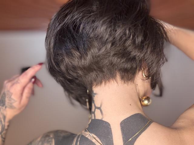 HannaBorbon's short, wavy black hair, tattoos on her neck and back, gold earrings, and hand with red nails, showing off her tattoos.