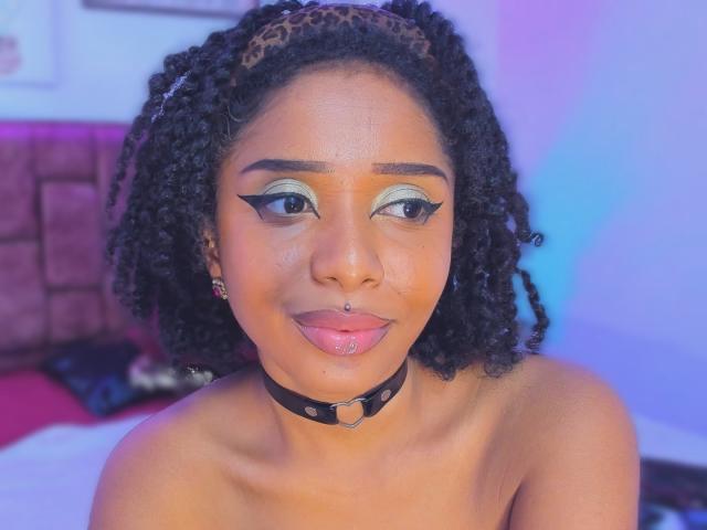 MeganWilliams with curly black hair, leopard headband, green eyeshadow, black eyeliner, pierced nose, pink lipstick, black choker, bare shoulders, looking at camera, smiling.