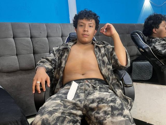 DreyFoster, un ragazzo robusto con i capelli ricci e neri, seduto su una sedia, con la camicia aperta che mostra un pancino morbido, indossa pantaloni e giacca mimetici, su sfondo blu..
