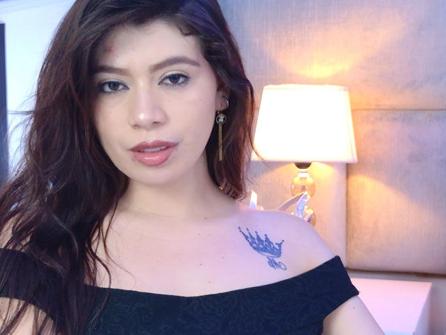 LindaMancini has long brown hair, light skin, and a small tattoo of a bird on her right shoulder. She's wearing an off-shoulder black top and a lamp is lit.