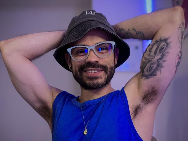 ThiagoKing con los brazos levantados, usando un gorro gris tipo cubo, una camiseta azul sin mangas, gafas y un collar, sonriendo, mostrando vello axilar, y tatuajes en su brazo derecho..