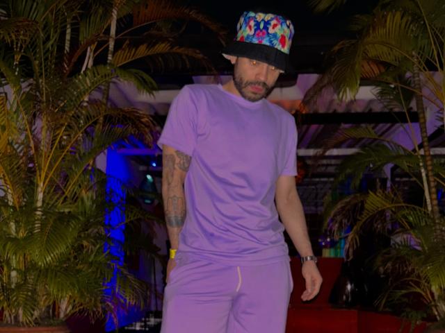 ThiagoKing wearing a purple t-shirt and pants, colorful hat, tattoos on right arm, dark beard, standing in front of palm trees, looking down with a serious expression.