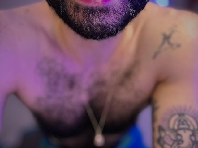 ThiagoKings behaarter Brust mit Tattoos, einem Kreuz auf der rechten Schulter und einer Halskette. Sein Bart ist sichtbar, und es gibt ein violettes Licht auf seinem Untergesicht..