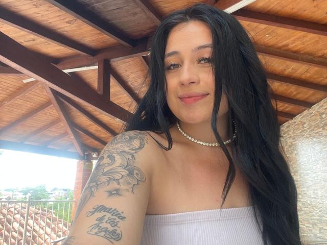 AnnyBentleii with long black hair, smiling, wearing a white tube top and pearl necklace, showing tattooed arm with text and floral design, standing under wooden roof.