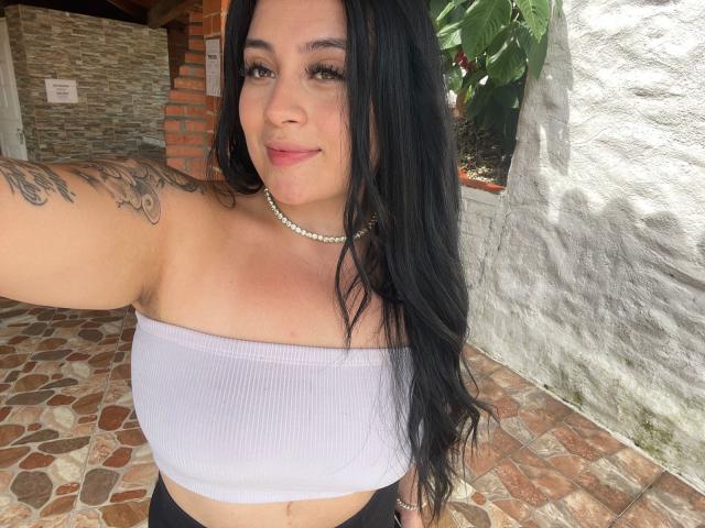 AnnyBentleii with long black hair, wearing a white tube top, showing her tits, tattoo on left arm, pearl necklace, smiling, standing on a brick patio.