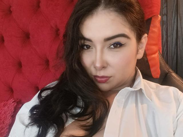 ValentinaCoba with long black hair, wearing a white shirt, looking at the camera with winged eyeliner, pink lipstick, and a pouty expression, against a red, tufted background.
