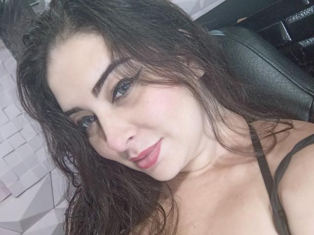 ValentinaCoba has wavy dark brown hair, blue eyes, and pink lipstick. She's wearing a black strap top, showing her small tits. She's smiling slightly, looking at the camera.