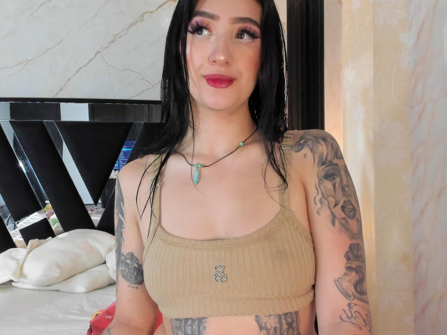 Tifany Diaz, con capelli neri, ombretto rosa e rossetto rosso, indossa un top corto beige a coste, mostrando tatuaggi sulle braccia e sul petto, e porta una collana verde..