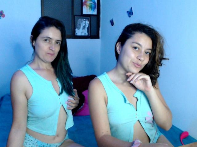 LunaVenus with dark green hair, wearing a teal, button-down top, exposing small tits, and light blue pants, sits on a bed, smiling, with one hand near her mouth.