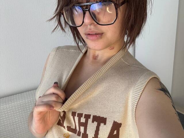 BarrieGromley with short brown hair, glasses, nose ring, and lip ring, wearing a beige sleeveless knit top, holding it with right hand, showing a small bruise on neck.