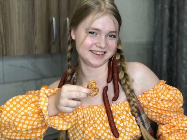 VeronaXXX – eine Frau mit geflochtenem, blondem Haar, die lächelt und ein Brezel hält. Sie trägt ein orangefarbenes Kleid mit Schulterfrei-Schnitt und weißen Mustern und befindet sich in einer Küche..