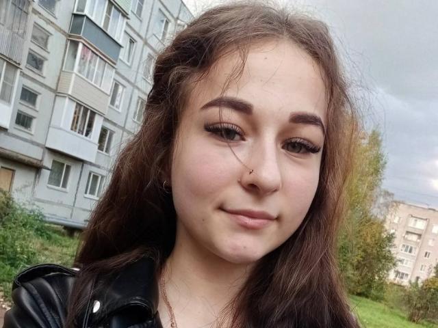 CherelleDelozier with long brown hair, small nose ring, and subtle smile, wearing black leather jacket, has slightly chubby cheeks and small tits, standing outdoors with greenery.