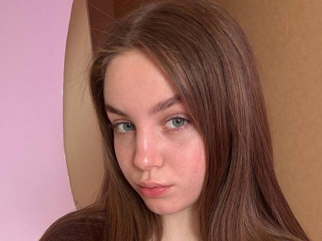 VellaDiehm a les cheveux bruns et lisses, des yeux verts, et une expression sérieuse avec des lèvres rosées légèrement retroussées. Elle a de petites taches de rousseur claires sur le nez et les joues..