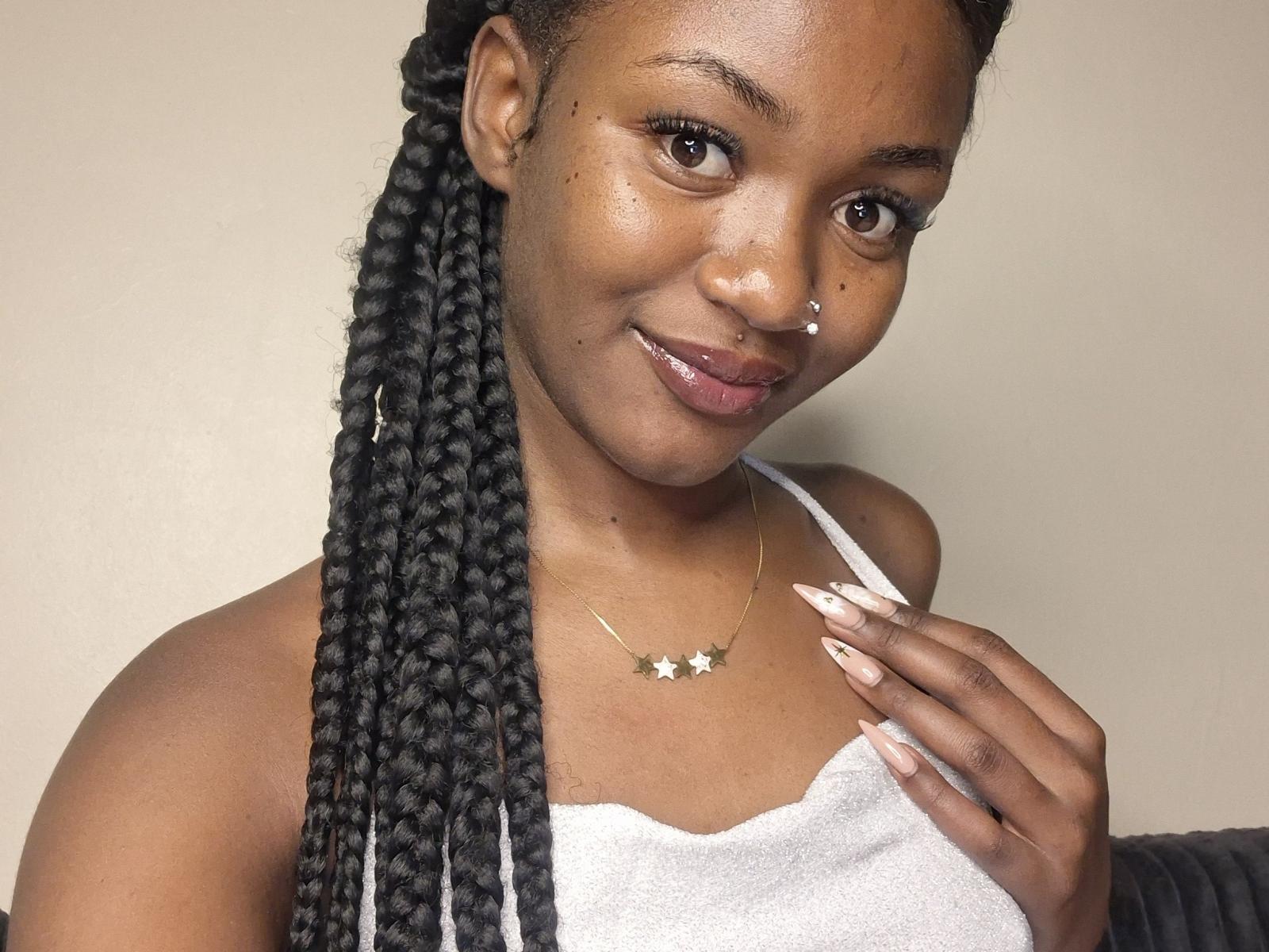 MissMilagros has dark brown skin, long black braids, a nose ring, and a smile, wearing a white tank top with a star necklace, and has long, manicured nails.