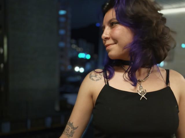 DarckVelvet with purple hair, black choker, and black top, showing off tattoos on shoulder and arm, smiling at camera, necklace with pentagram, city lights in background.