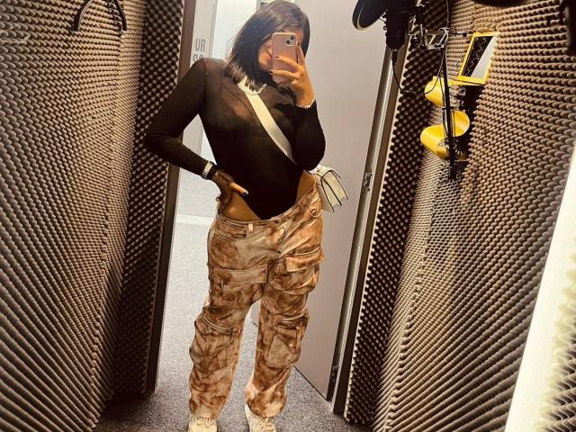 ValerySilva with medium tits, wearing a sheer black top, camo pants, and white sneakers, taking a mirror selfie in a room with textured walls and a yellow light.
