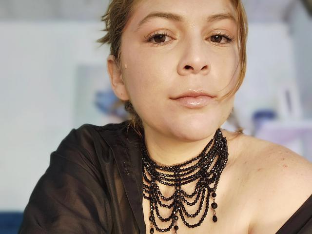 MoonXVelvet, com pele clara, olhos castanhos e lábios brilhantes, usando um colar preto com contas e uma blusa preta transparente, tem um leve sorriso e seu cabelo está preso para trás..