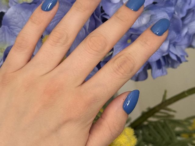 KarKutie's hand with blue, shiny, almond-shaped nails, slightly spread fingers, pale skin, and a few small wrinkles, set against a blurred purple flower.