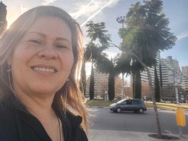 CoupleBomb has a chubby face, light brown hair, and is smiling with small tits. She's wearing a black top, standing outside in a sunny urban area.